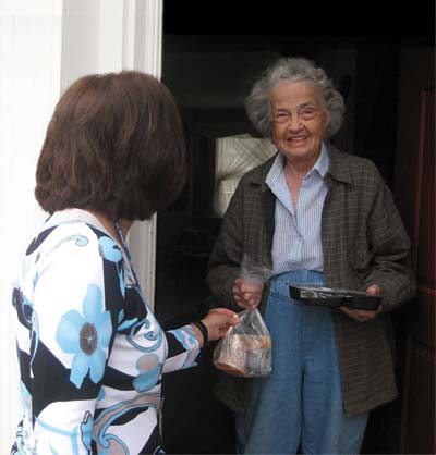 Older woman standing in doorway smiling as brunette woman hands her food